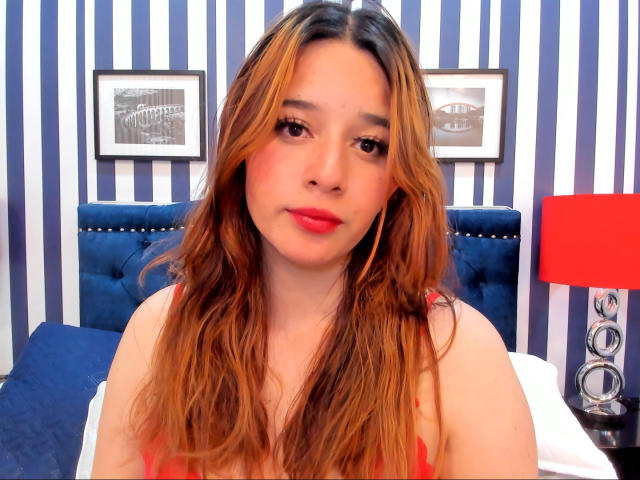 DaniVelasquez with long, wavy, brown-to-blonde ombre hair, red lipstick, and a cute face, wearing a red top, sitting in front of a blue, tufted headboard.