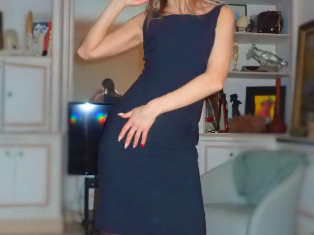 KatarinaDamo in a tight black dress, hand on her ass, other arm raised, blonde hair, red nails, standing in a room with a TV and white furniture.