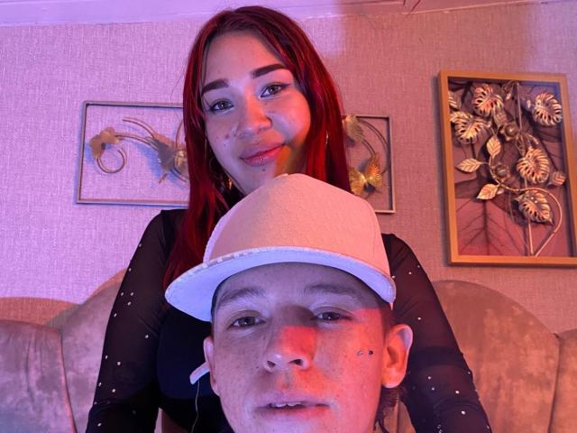 PhoenixEvelyn with red hair and makeup, black top, leans over a guy with a white hat, nose piercing, and earbuds, both looking at the camera.