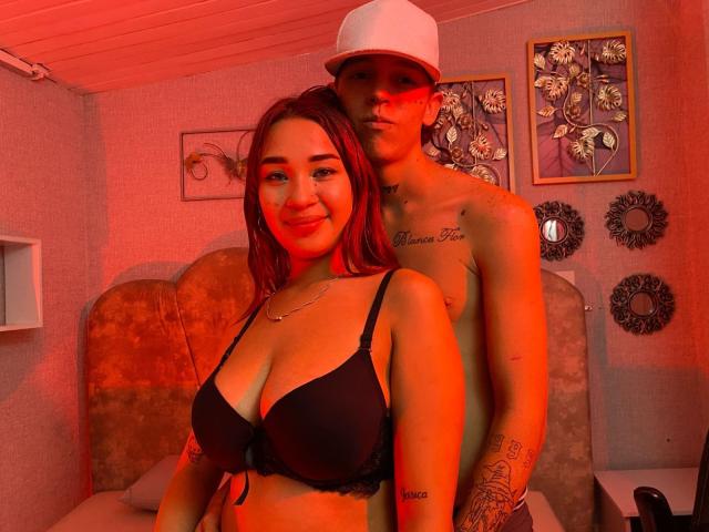 PhoenixEvelyn in reggiseno nero, in piedi di fronte a un ragazzo senza maglietta con un berretto bianco, illuminazione rossa, tatuaggi sul suo braccio e sul petto, entrambi sorridenti, in una stanza accogliente..