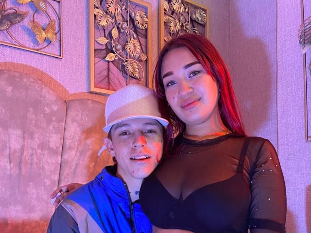 PhoenixEvelyn with red hair, black sheer top, and tits, and a dude in a white hat and blue jacket, smiling in a purple-lit room. PhoenixEvelyn's hand on dude's shoulder.