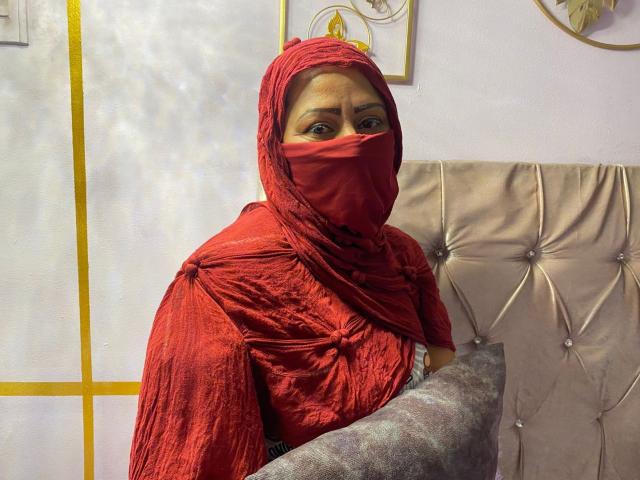 AmiraAb, wearing a red veil and matching dress, holds a gray pillow, with her brown eyes peering through the fabric, has a medium brown skin tone, and a serious expression.