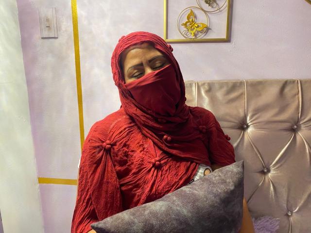 AmiraAb, wearing a red, textured hijab and matching mask, holds a gray pillow. Her eyes are half-closed, and she's sitting in front of a tufted, beige headboard.