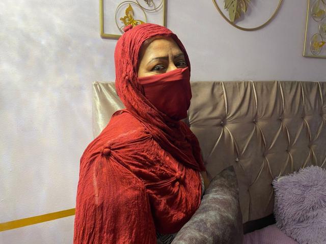 AmiraAb has red cloth covering her head and mouth, with her tits slightly visible, wearing a red dress, sitting on a gold-tufted couch, looking up with a worried expression.