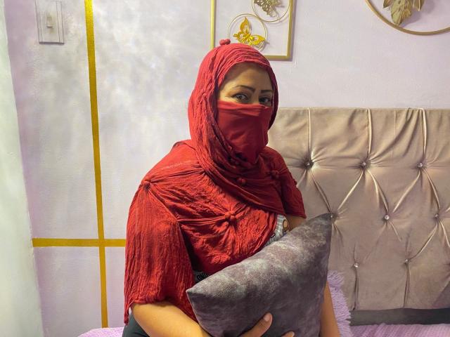 AmiraAb with red hijab and matching mask, holding a grey pillow, wearing a textured red top, sitting on a tufted beige bed, looking slightly to the side.