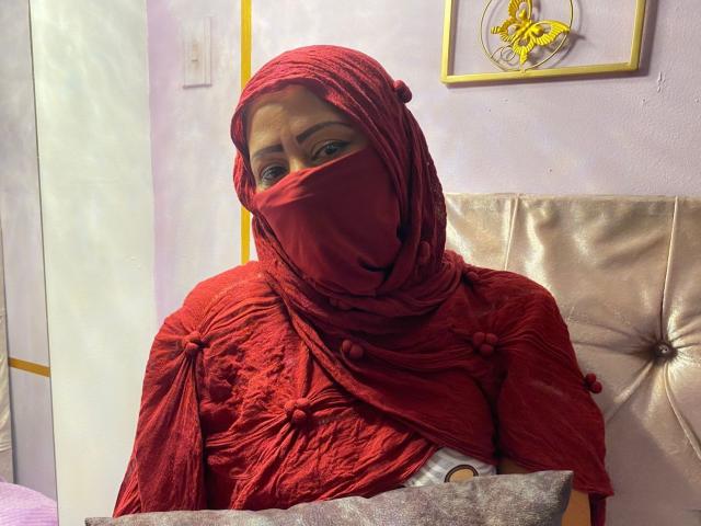 AmiraAb with red hijab and mask, wearing textured red robe, sitting on silver chair, holding gray pillow, looking slightly to the side, purple wall in background.