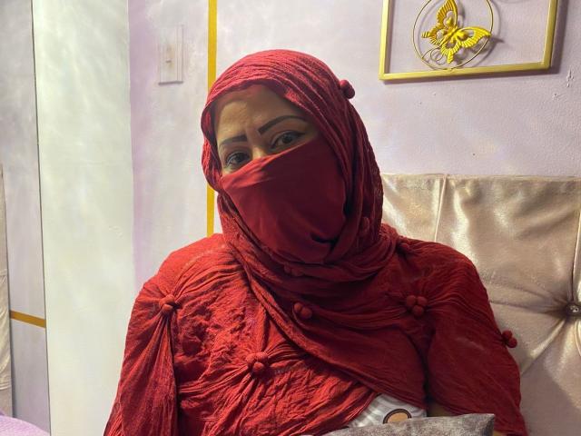 AmiraAb with red hijab and matching red dress, covering her nose and tits, sitting on a beige chair, looking at the camera, in a softly lit room.