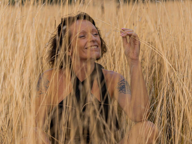 ZoraEnn-Official-hot with wavy brown hair, smiling, holding a straw, tattoo on right arm, wearing black sleeveless top, sitting in golden wheat field. Her tits are partially visible.