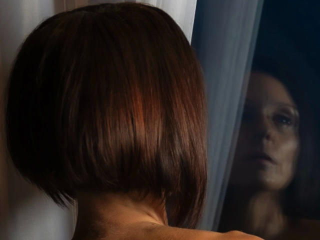 ZoraEnn-Official-hot with shiny brown bob, looking in mirror. Her reflection shows serious face, slight shadow on right side. Her butt is partially visible, upper back exposed.