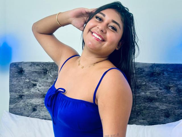 AmmyRoux with dark hair, smiling, wearing a blue dress, hand in hair, gold bracelet, necklace, and earrings, sitting in front of a tufted grey headboard.