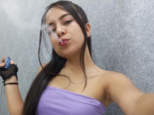 JessyLenon with long black hair, pouty lips, purple strapless top, black fingerless glove, and silver necklace, holding a blue object, winking. Smoky breath near her face.