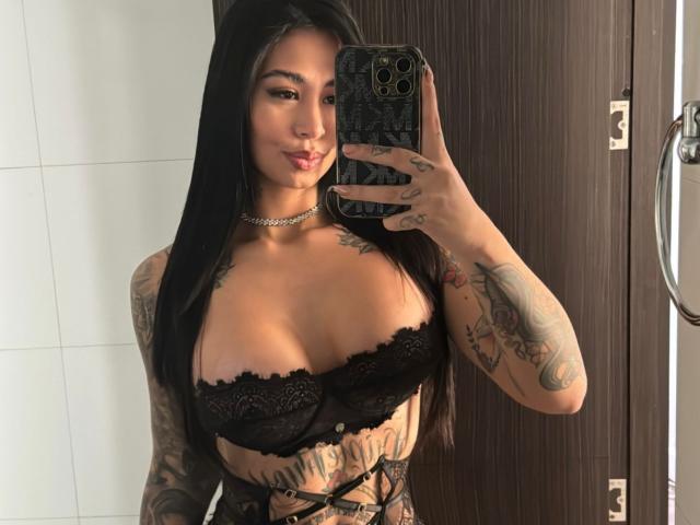 ValentinnaSquirt, con cabello negro largo, vistiendo un sujetador de encaje negro, mostrando sus grandes senos, tatuajes en los brazos y el pecho, sosteniendo un teléfono, selfie frente al espejo, collar tipo gargantilla, expresión de labios fruncidos, puerta marrón..