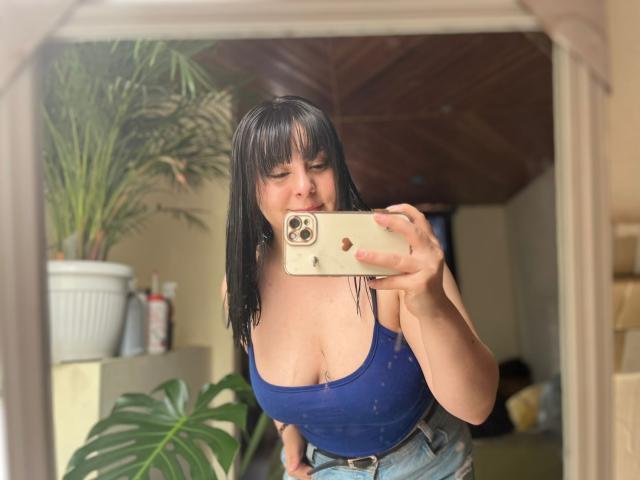EmaJames with black hair, wearing a blue tank top, showing big tits, taking a mirror selfie, holding a gold iPhone, standing in a room with plants.