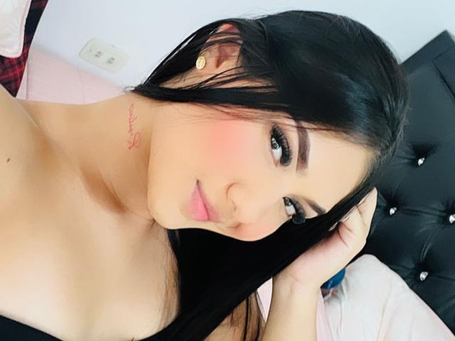 VioletJhons, com cabelo preto, blush rosa e rosto fofo, tocando o cabelo, mostrando seus ombros nus e uma pequena tatuagem vermelha no pescoço. Seus seios estão parcialmente visíveis..
