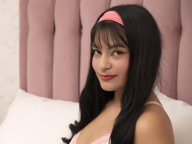 JulietaRizzo with long black hair, pink headband, and pink lace bra, smiling softly, showing her cute face and small tits, sitting on a bed with white pillows.