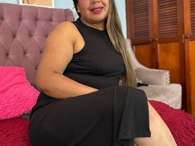 EmilyGhrey, a plus-sized woman with tan skin and long brown hair, sits on a bed in a black dress, showing her tits and smiling with pink lipstick.
