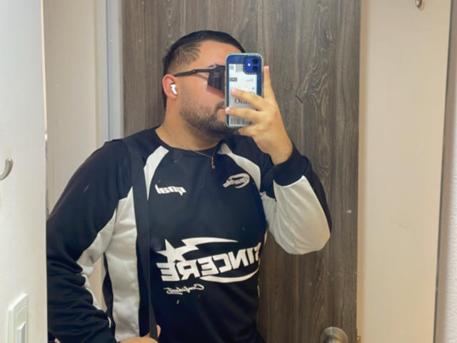 GaelB mit kurzen, dunklen Haaren, Bart und Brille, trägt ein schwarz-weißes Sportshirt und macht ein Selfie mit einem blauen Handy, wobei weiße Kopfhörer im Ohr sind..