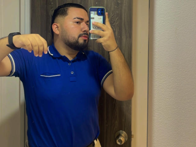 GaelB com barba, vestindo uma camisa polo azul, tirando uma selfie no espelho, segurando o celular com uma mão, levantando a outra, mostrando uma pulseira preta..