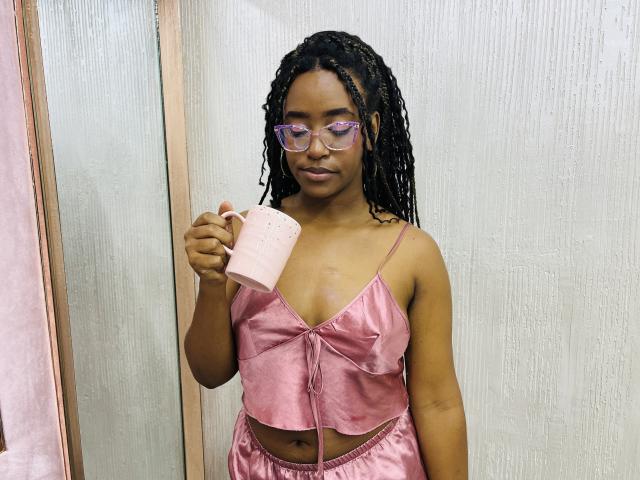 NaomiReyes with braided hair, pink glasses, and pink satin crop top, holding a pink mug, showing her small tits and a belly button tattoo.