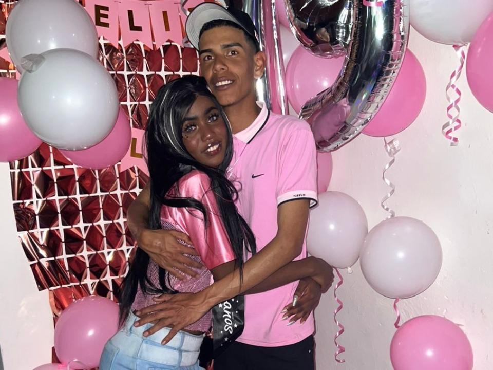 AishaAndM, con la pelle scura, i lunghi capelli neri e piccoli seni, abbraccia un ragazzo alto dalla pelle scura che indossa una maglietta rosa, circondati da palloncini rosa e bianchi. Lui sta sorridendo..