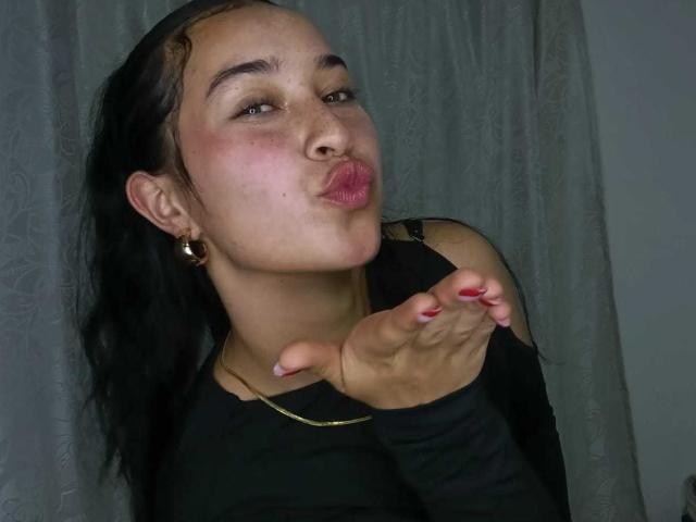 SolOrtega infla seus lábios rosados, fazendo uma careta de beijo. Ela tem cabelo escuro, brincos de argola e unhas vermelhas, vestindo uma blusa preta com recortes nos ombros..