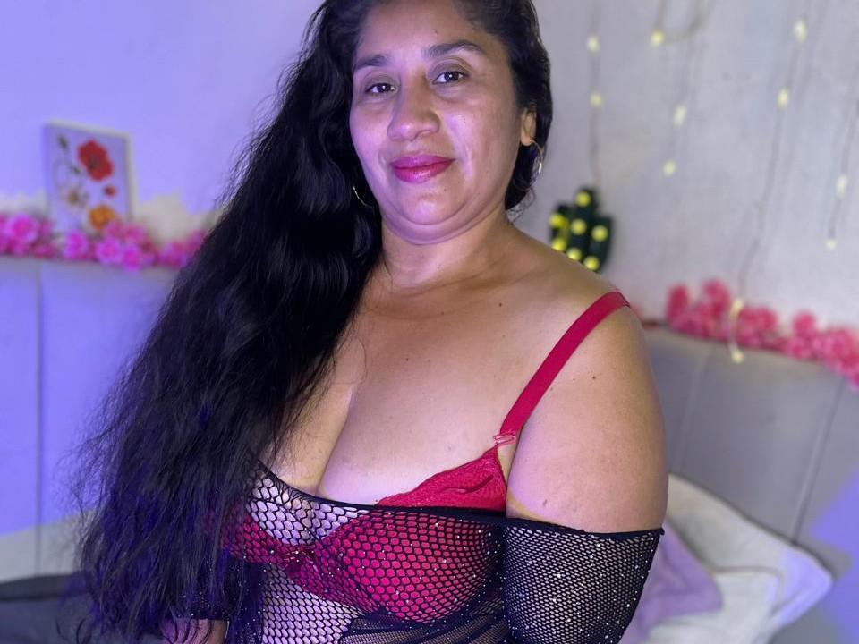 MilfLatine with long black hair, red bra, and black fishnet sleeves, showing her tits, smiling, pink lipstick, and hoop earrings, in a bedroom with soft lights.
