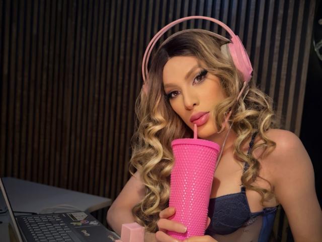 Putinna with pink headphones, curly blonde hair, and pink straw in a cup, sucking through it, holding laptop, wearing blue bra, showing her tits, in a cozy setting.