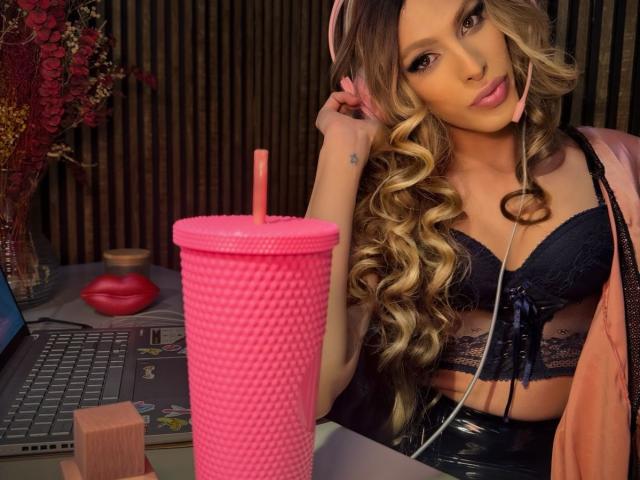 Putinna with curly blonde hair, pink headphones, black lace bra, and pink jacket, holding a pink textured drink with a straw, next to a laptop and red lips.