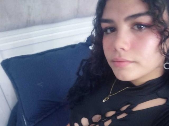 BrianaCleython with curly black hair, wearing a black, ripped shirt, gold necklace, and pink eyeliner, looking at the camera with a neutral expression. Her tits are visible through the shirt.