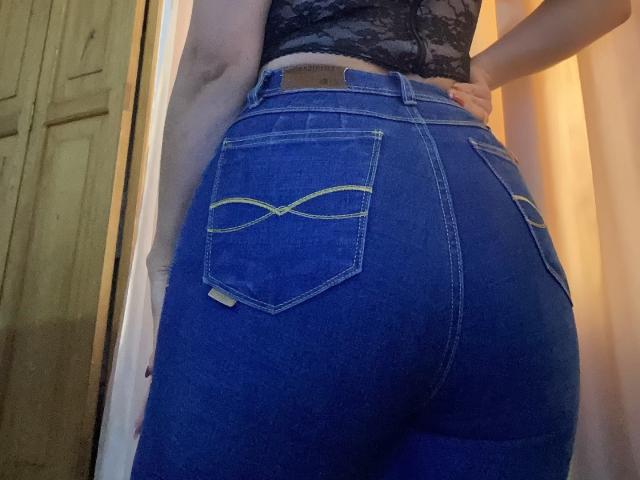 MiaDuboisX's tight blue jeans show off her butt, featuring yellow double-stitched pockets. She's wearing a black lace top, with her hand on her hip.