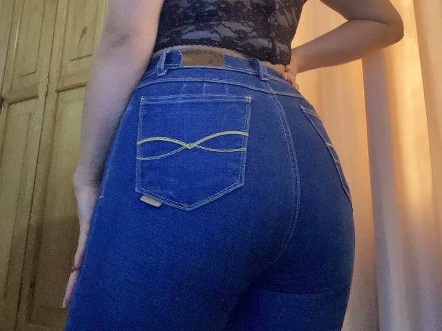 MiaDuboisX's ass in tight blue jeans with yellow double-arch pockets, lace top, hand on hip, standing, photo shows her back, slightly bent, in dim light.