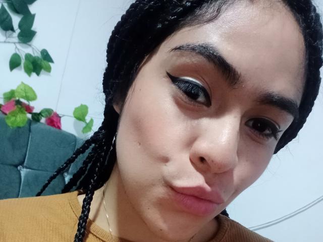StellaBeckker with puffy lips, black braided hair, thick eyebrows, and black winged eyeliner, wearing a mustard top, has a cute, slightly mischievous facial expression.