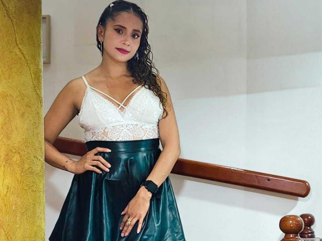 EliBaker with curly brown hair, white lace top, and green leather skirt, standing, hand on hip, black wristband, red lipstick, yellow wall, wooden railing, smiling.
