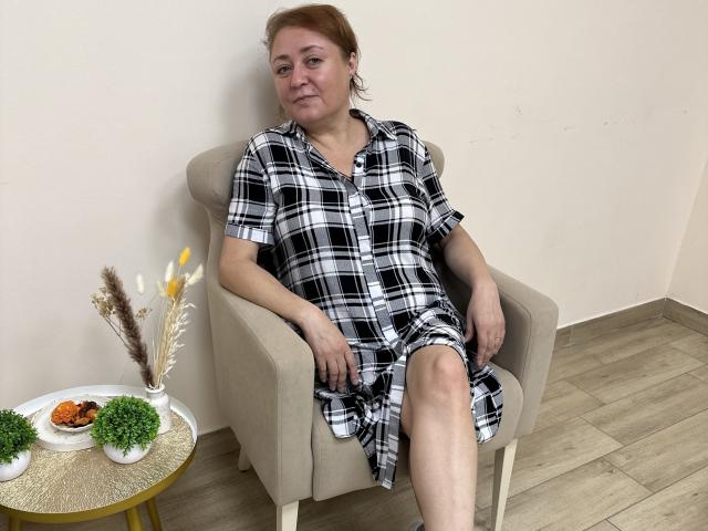 VioletHarmon, middle-aged woman with red hair, wearing a black and white plaid dress, sitting in a beige chair, slightly smiling, with her legs crossed. Small table with plants and snacks.