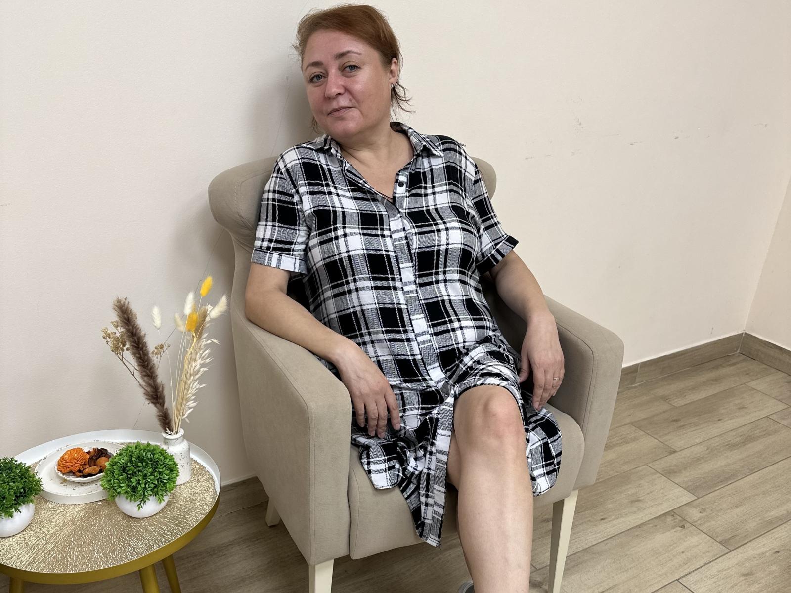 VioletHarmon, middle-aged woman with red hair, wearing a black and white plaid dress, sitting in a beige chair, slightly smiling, with her legs crossed. Small table with plants and snacks.