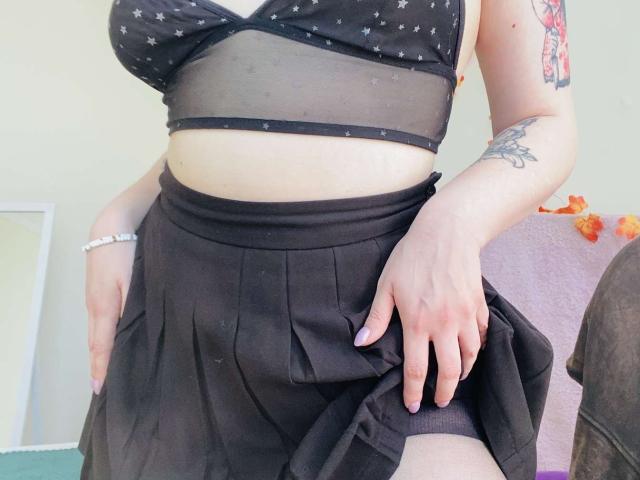 KarKutie's boobs in a sheer black star-patterned bralette, black pleated skirt, left hand on her thigh, right hand on her skirt, visible tattoos on her right arm.