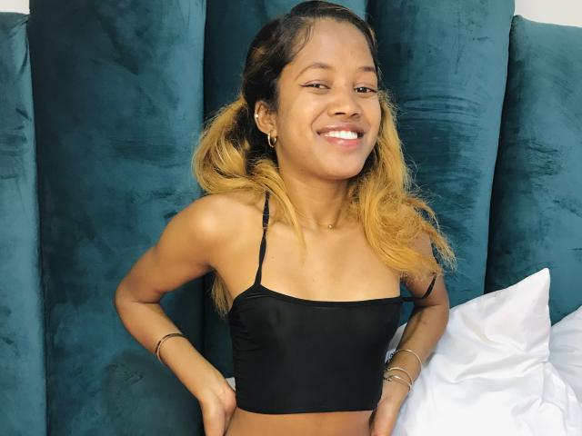 SaynahSisi with a cute smile, black strapless top, tan skin, blonde and brown hair, hands on hips, wearing gold bracelets, blue velvet headboard, white pillow in background.