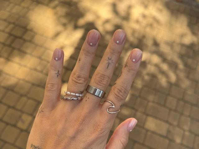 Tsukerberg's hand with pinkish-nude nails, star tattoos, and three rings (metal, silver, heart-shaped) on a sunlit brick surface. Boobs not visible.