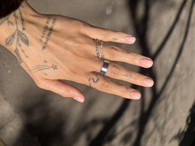 Tsukerbergs hand, met roze nagels, een zilveren ring en een zwarte metalen ring, met tatoeages van bladeren, sterren en letters, op een grijze achtergrond..