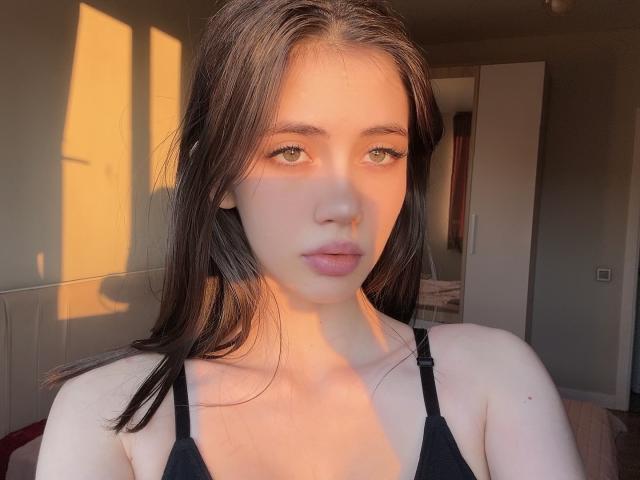 SweetyVikky with long brown hair, green eyes, and pink lips, wearing a black bra, bathed in warm sunlight. She's looking straight at the camera, her face slightly flushed.