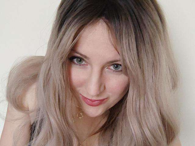 EuniceKiss has blonde, slightly wavy hair, green eyes, and a cute smile with pink lipstick. She's leaning forward, showing a small gold necklace against a white background.