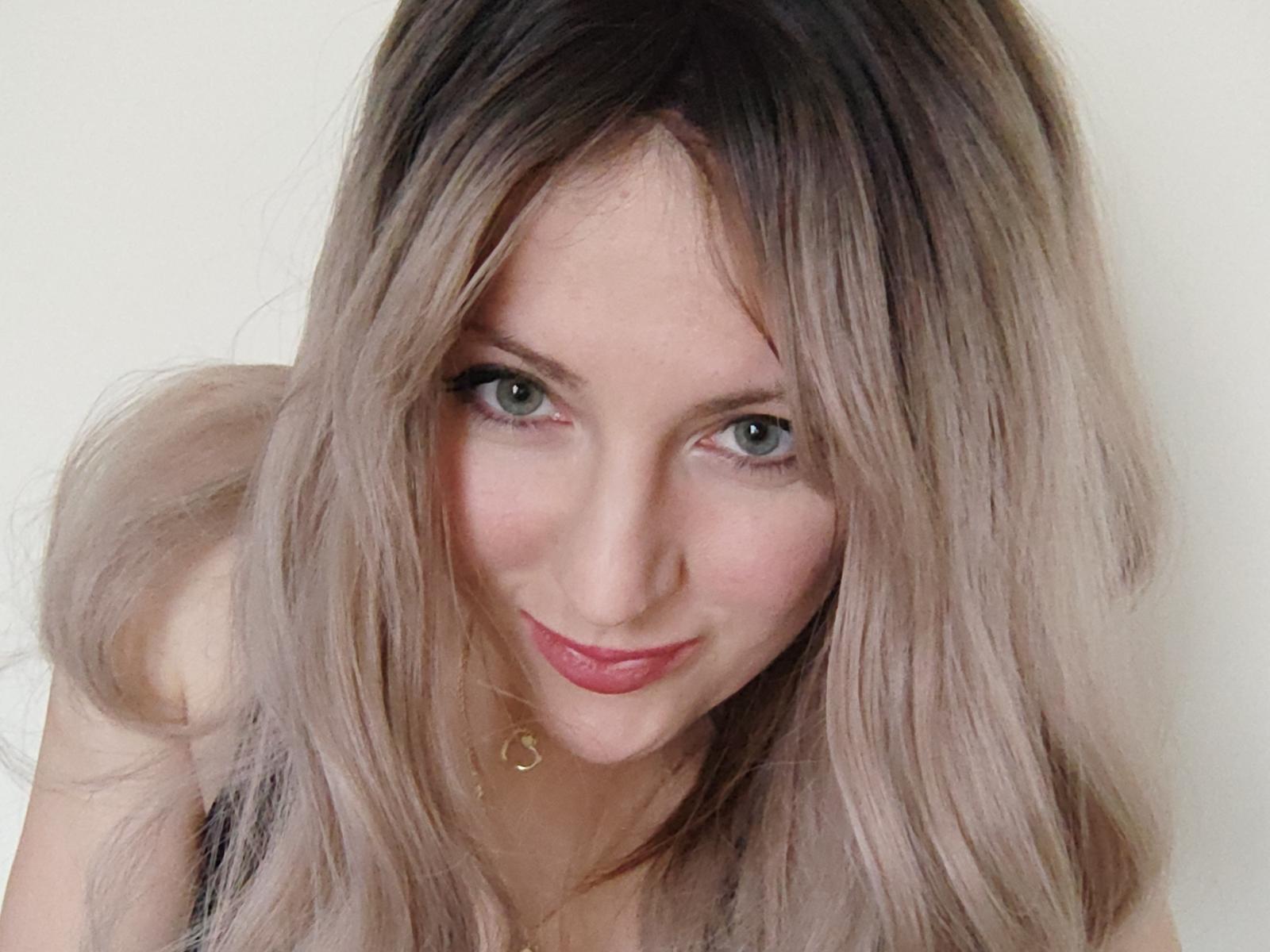 EuniceKiss has blonde, slightly wavy hair, green eyes, and a cute smile with pink lipstick. She's leaning forward, showing a small gold necklace against a white background.