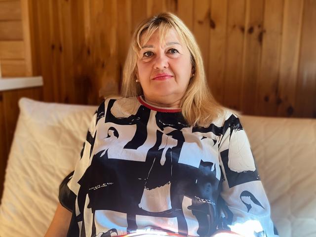 EmmaEllison with blonde hair, red lipstick, black-and-white abstract pattern shirt, sitting on white pillows, wood-paneled wall background, smiling slightly, medium build, cute, relaxed.