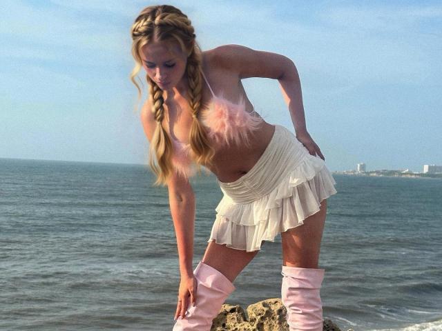 CamiiLaa with long blonde braids, topless, wearing a pink feathered bra, white pleated skirt, and pink thigh-high boots, leaning forward by the ocean. Her tits are visible.