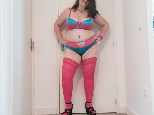 LiliChattePoilue stands in pink and blue lingerie, including pink stockings and high heels, showing her big tits, belly, and curvy ass, against a white door.