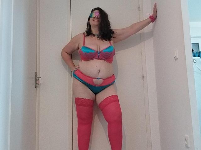 LiliChattePoilue stands in a doorway, wearing pink and blue lingerie with red thigh-highs. She has a curvy, plus-sized body with medium tits, a belly button piercing.