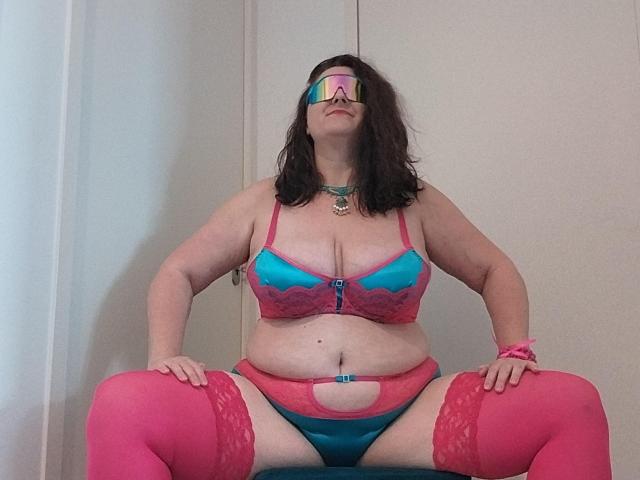LiliChattePoilue with medium tits, pink and blue lingerie, pink thigh-highs, and a reflective visor, sitting with legs apart, showing a cut-out in her panties.