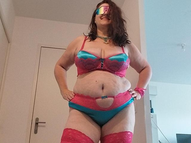 LiliChattePoilue, plus-sized with pale skin, wearing pink and blue lace lingerie, red thigh-highs, and rainbow glasses, standing in a room, hands on hips, smiling.