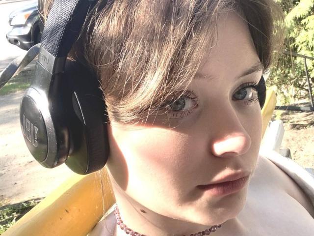 NikolKitt has light brown hair, blue eyes, and is wearing black Razer headphones with a serious expression, sunlight shining on her face, and a pink necklace.