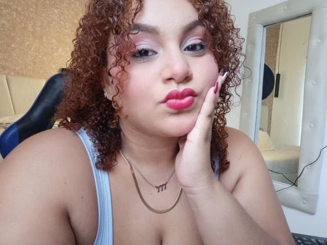 YeraCarey has curly brown hair, pink lipstick, and is pouting with her finger on her cheek. She's wearing a silver necklace and a blue tank top.